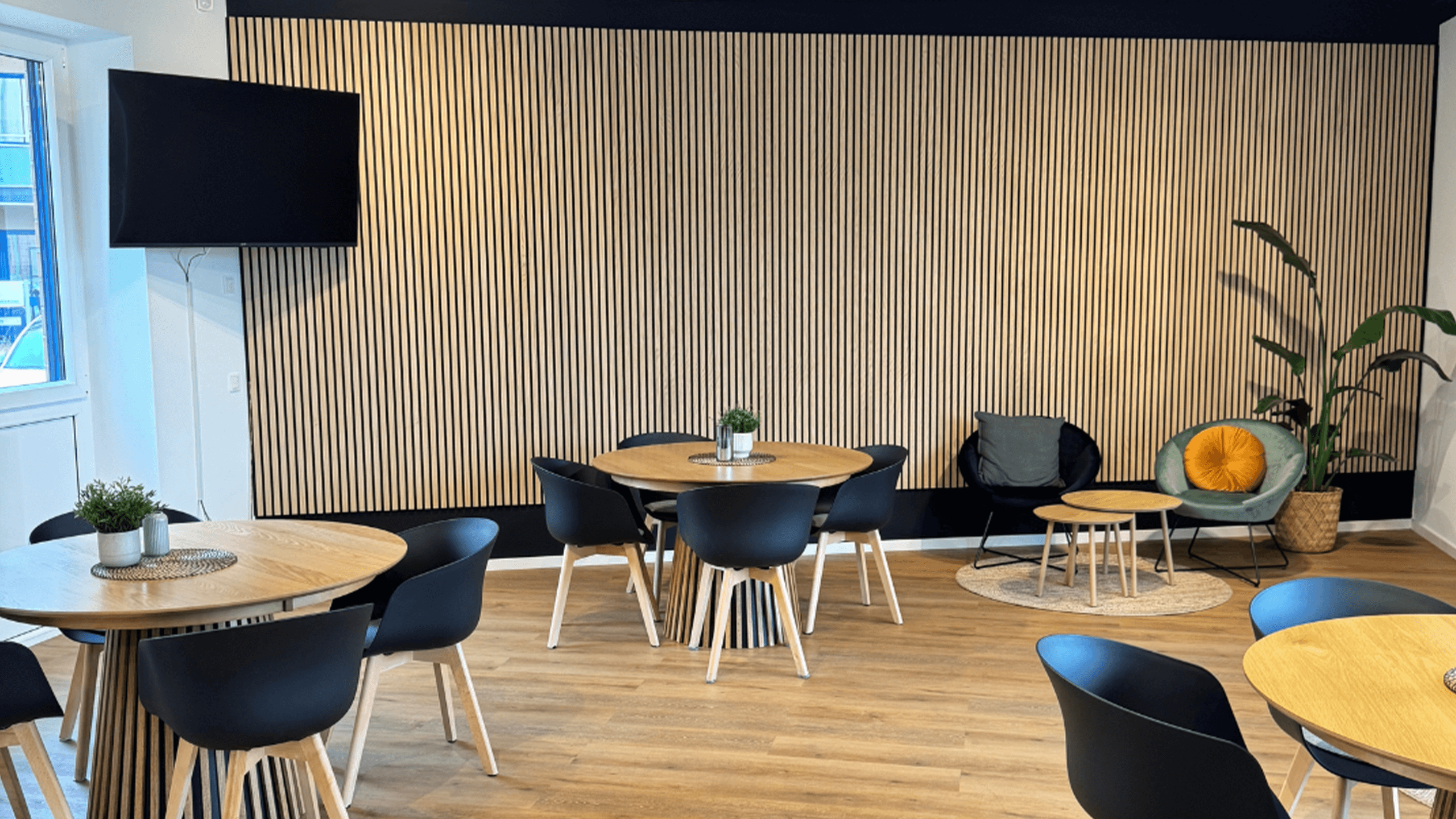 Moderner Pausenraum mit runden Holztischen, schwarzen Stühlen, gemütlichen Sesseln, Pflanzen und einem großen Fernseher an der Wand.